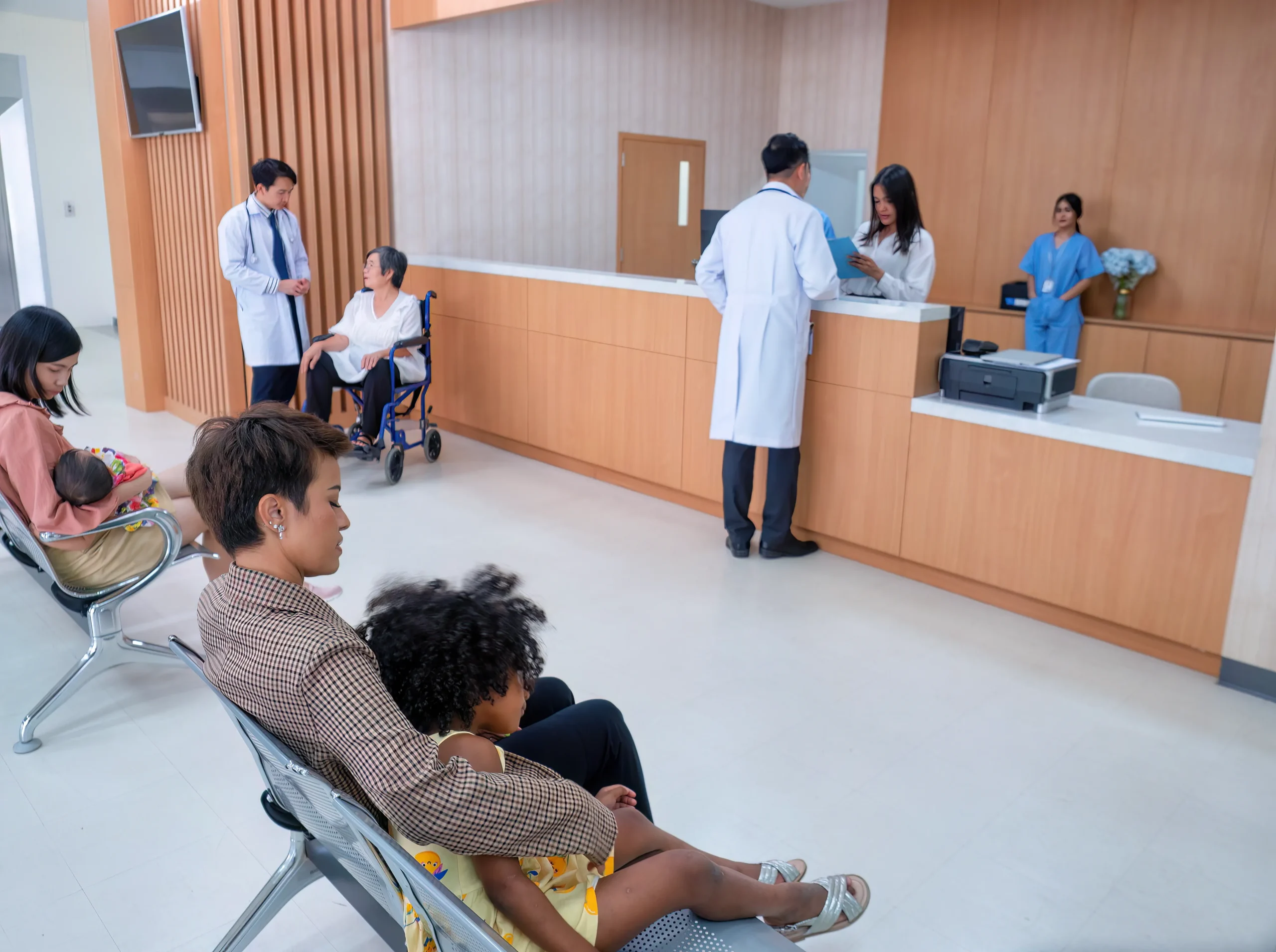 People are sitting in a hospital waiting area; a mother with a baby, and a child on a woman's lap. Doctors converse with patients at the reception.
