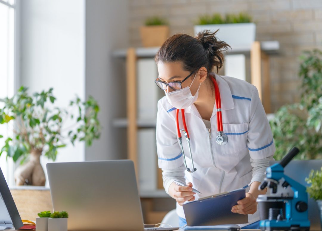 A female doctor in a white coat, mask, and red stethoscope reviews notes on a clipboard while standing in an office with plants and a microscope.