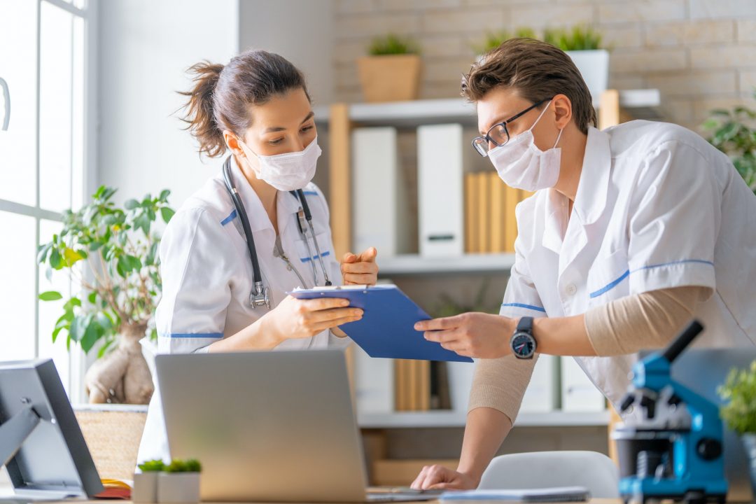 Healthcare professionals in masks discuss a blue clipboard in a bright office. A laptop and microscope are on the desk, suggesting a medical setting.